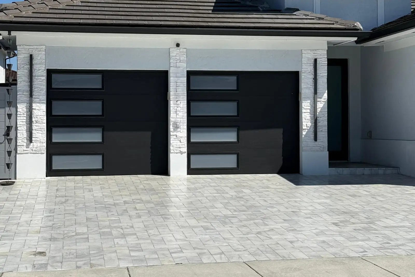 Two black garage doors with frosted window strips on a modern white home with stone-clad pillars and a paver driveway