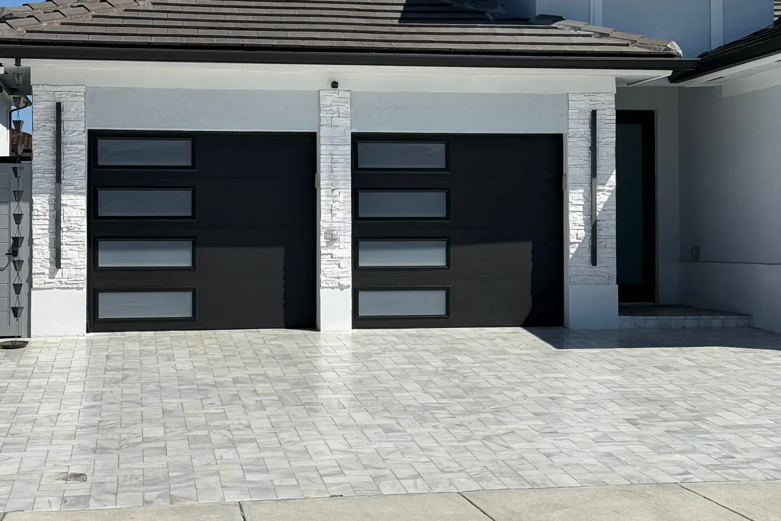 Two black garage doors with frosted window strips on a modern white home with stone-clad pillars and a paver driveway