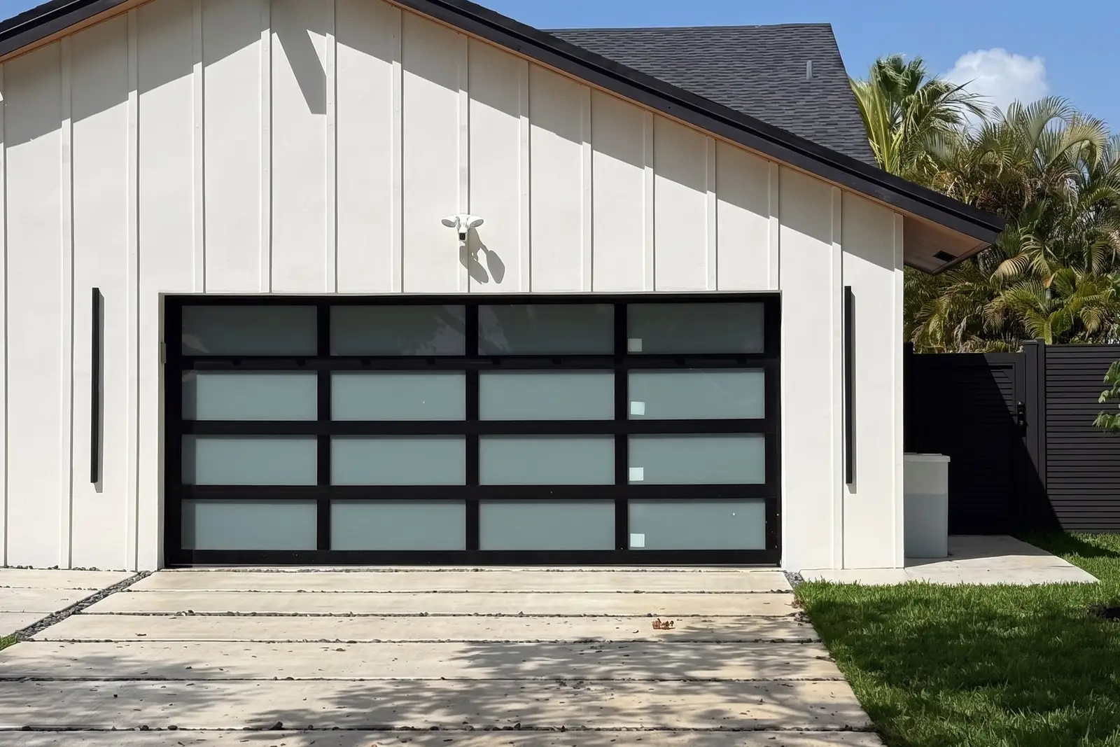 White board-and-batten house with a black-framed frosted-glass garage door, palm trees and a blue sky
