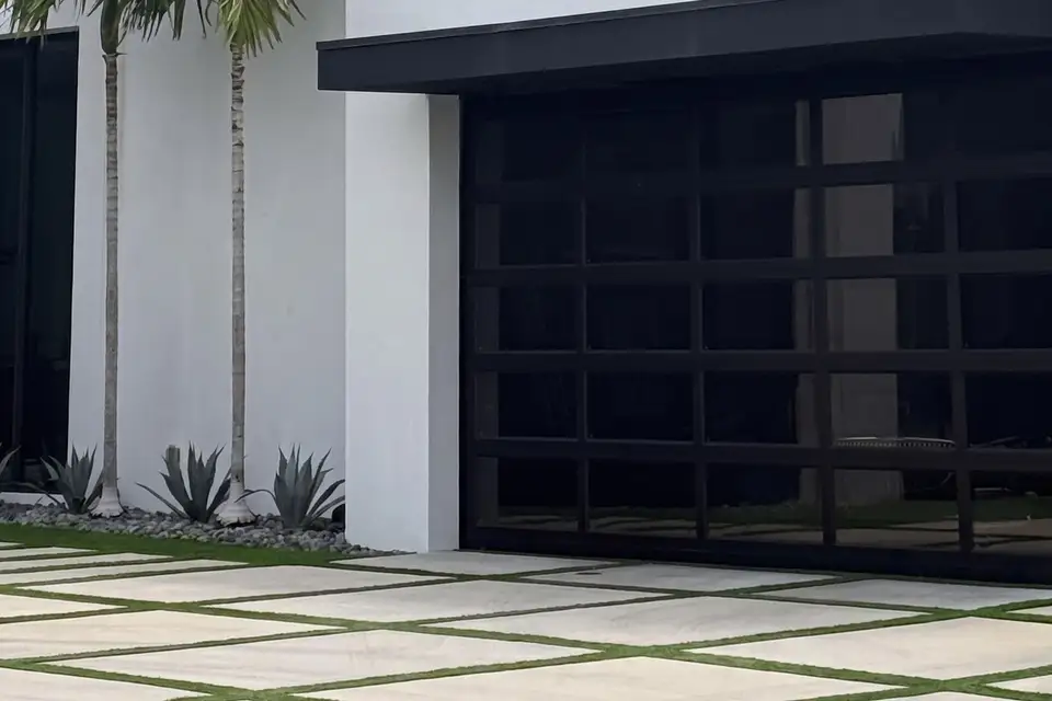 Black full-view glass garage door on a modern white home with palms, agaves and a grass-joint driveway