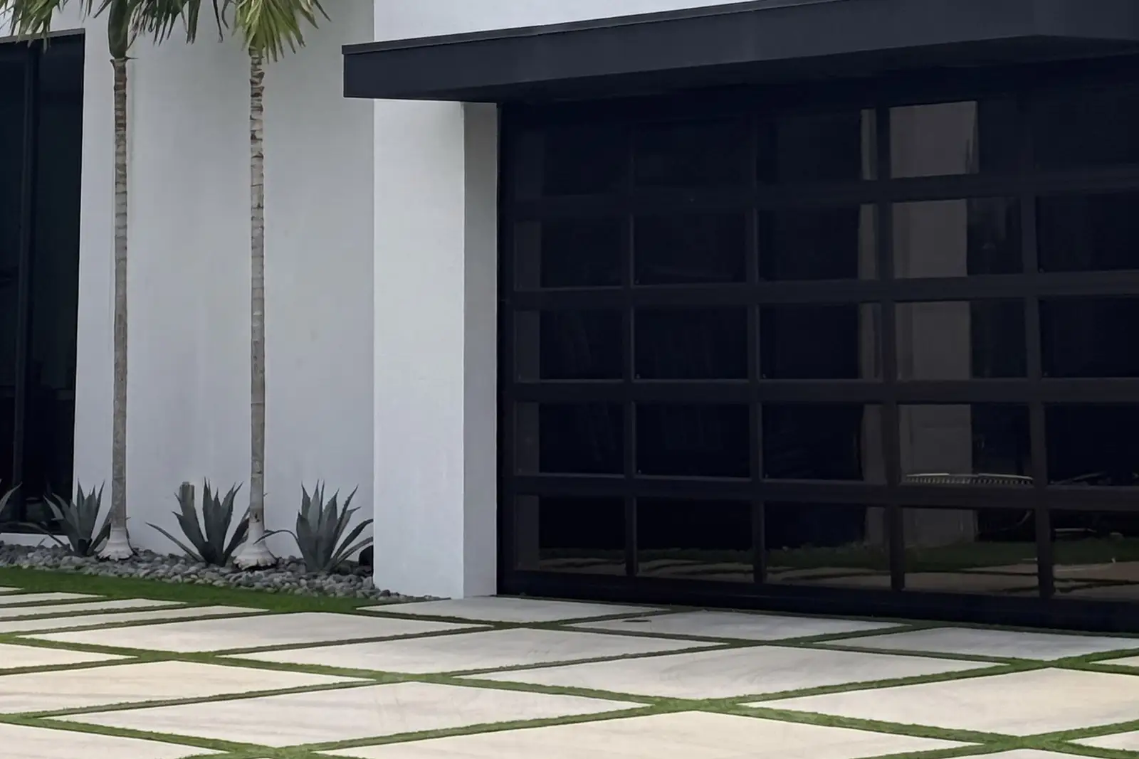 Black full-view glass garage door on a modern white home with palms, agaves and a grass-joint driveway